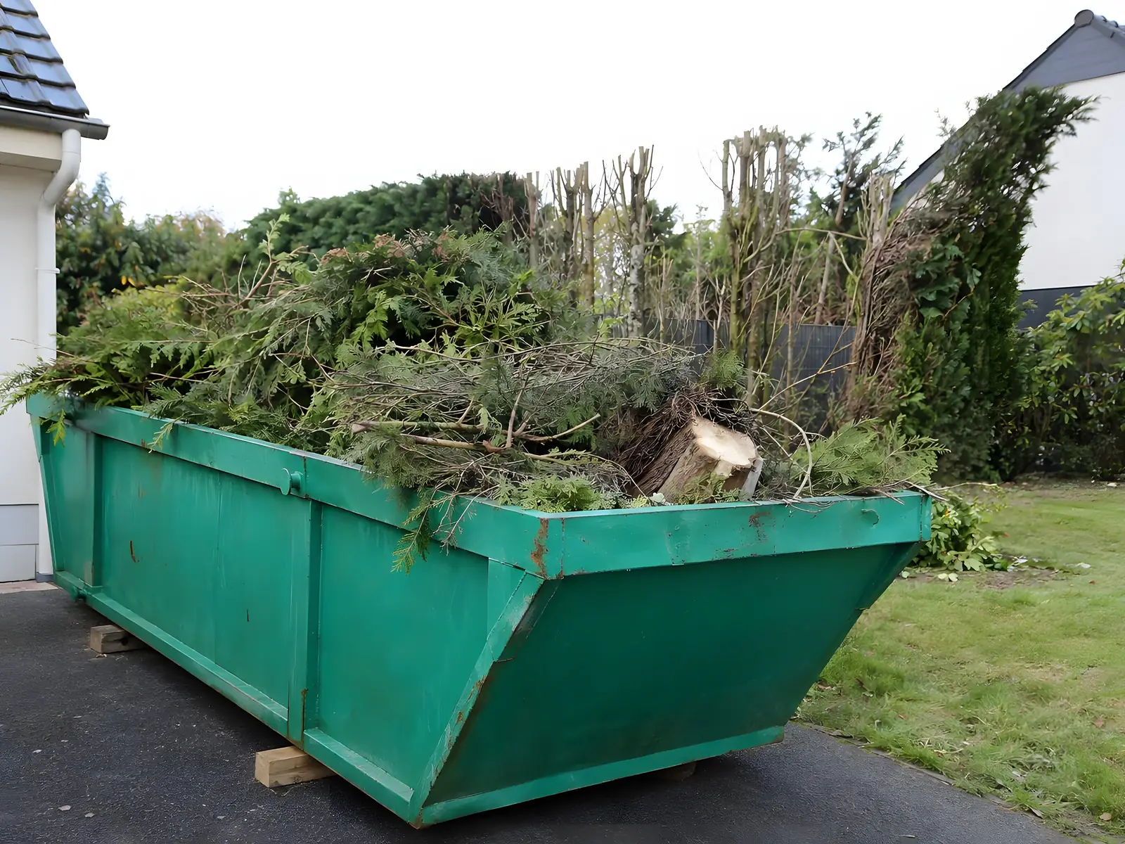 Benne verte remplie de branchages et de déchets de taille, posée au bord d’une pelouse.