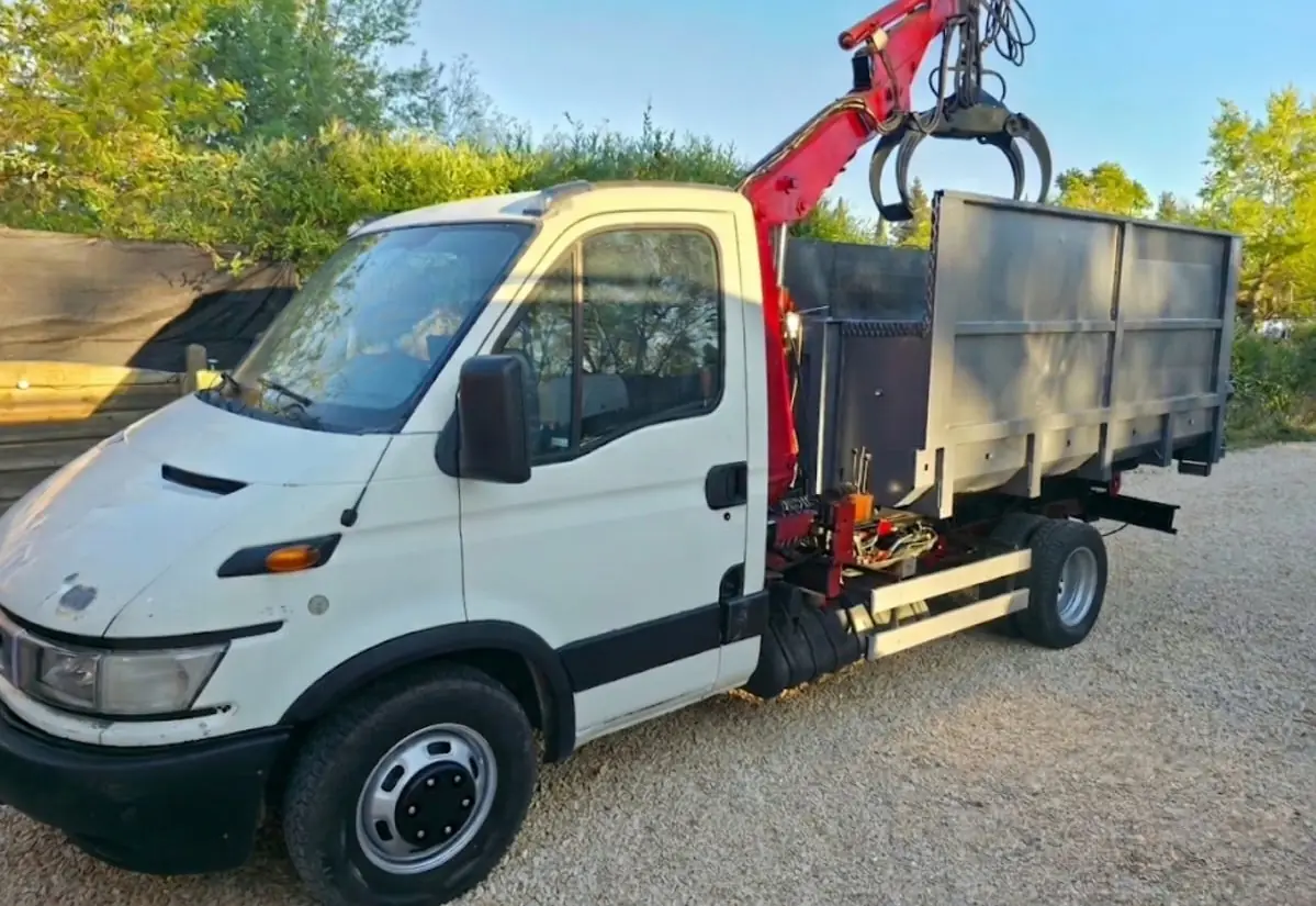 Le camion blanc de RJ Benne vu de trois quarts avant : grue rouge repliée, grappin au repos au-dessus de sa benne grise, sur du gravier.