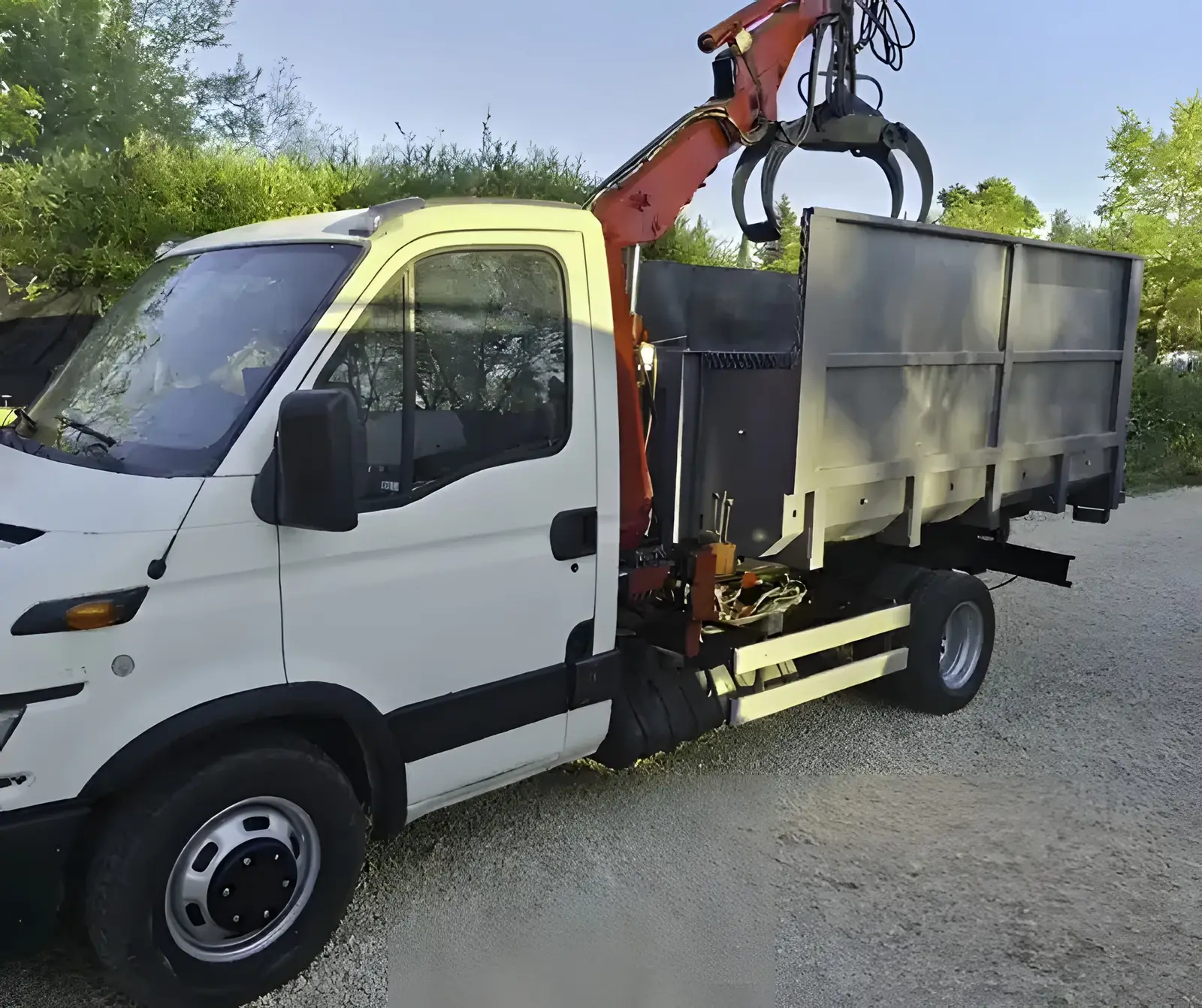 Le camion blanc de RJ Benne, grue orange repliée et grappin au repos au-dessus de sa benne grise, garé sur du gravier.