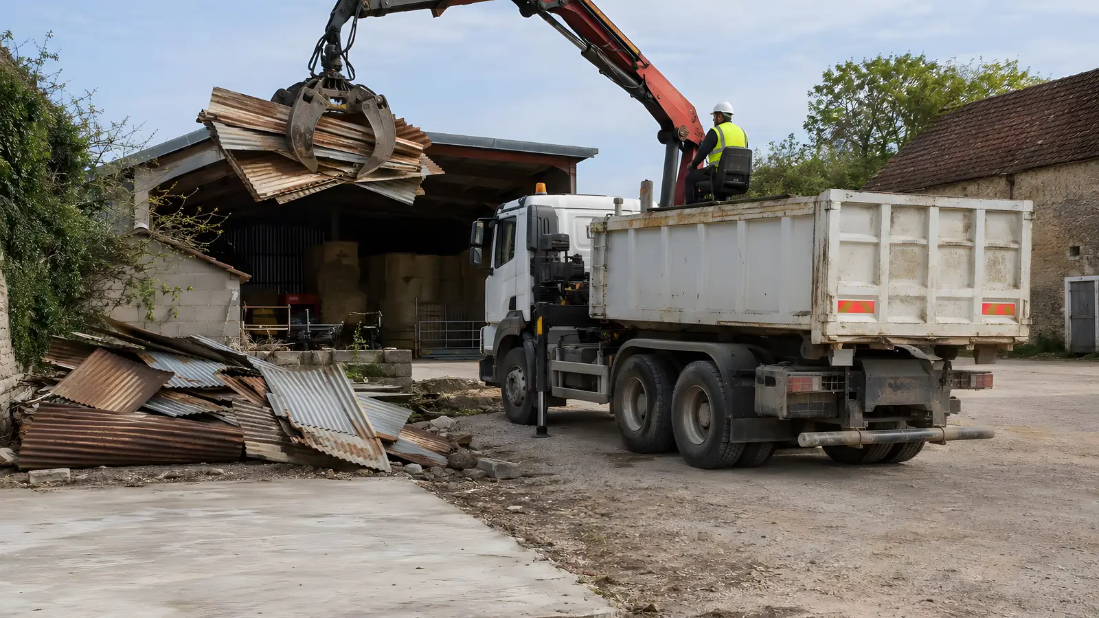 Camion grue dans une cour de ferme : depuis son siège, l’opérateur soulève au grappin des tôles ondulées rouillées, tirées du tas d’un abri démonté, pour les charger dans la benne.