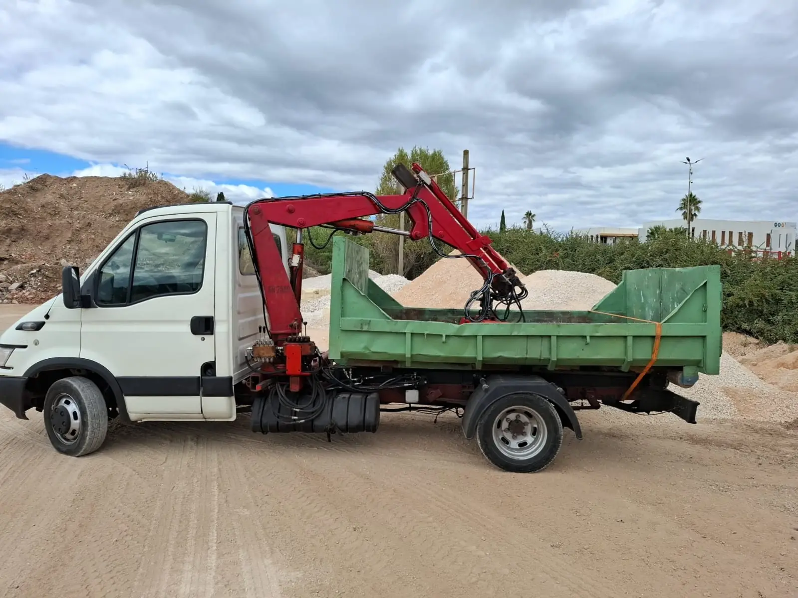 Le camion de RJ Benne vu de côté, grue rouge repliée dans sa benne verte vide, dans une cour de matériaux bordée de tas de sable et de gravier.