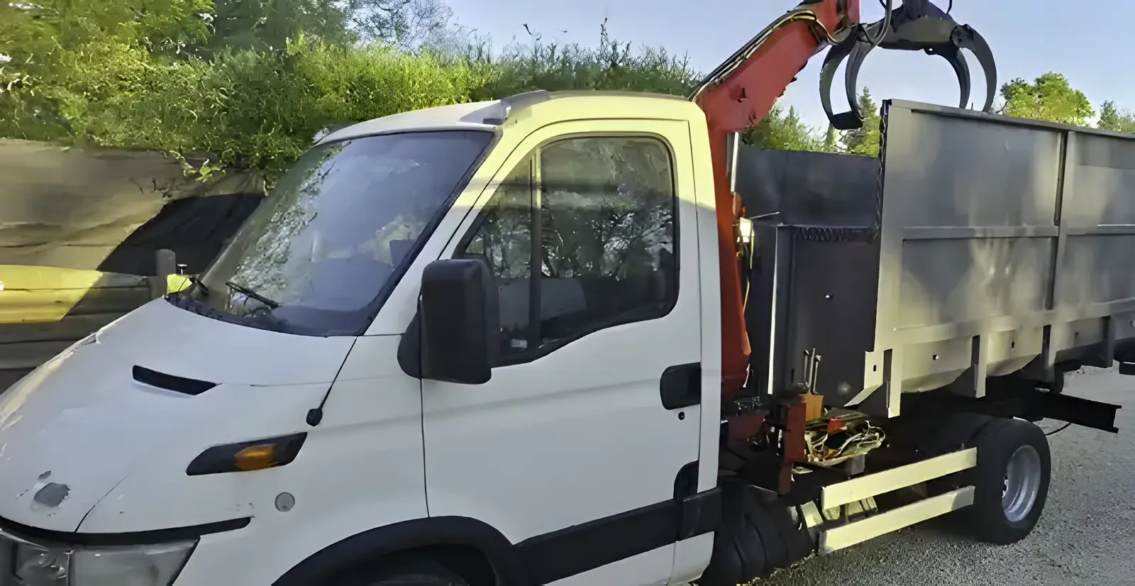 Le même camion blanc à grue orange et grappin, vu de trois quarts avant, sa benne grise à l’arrière.