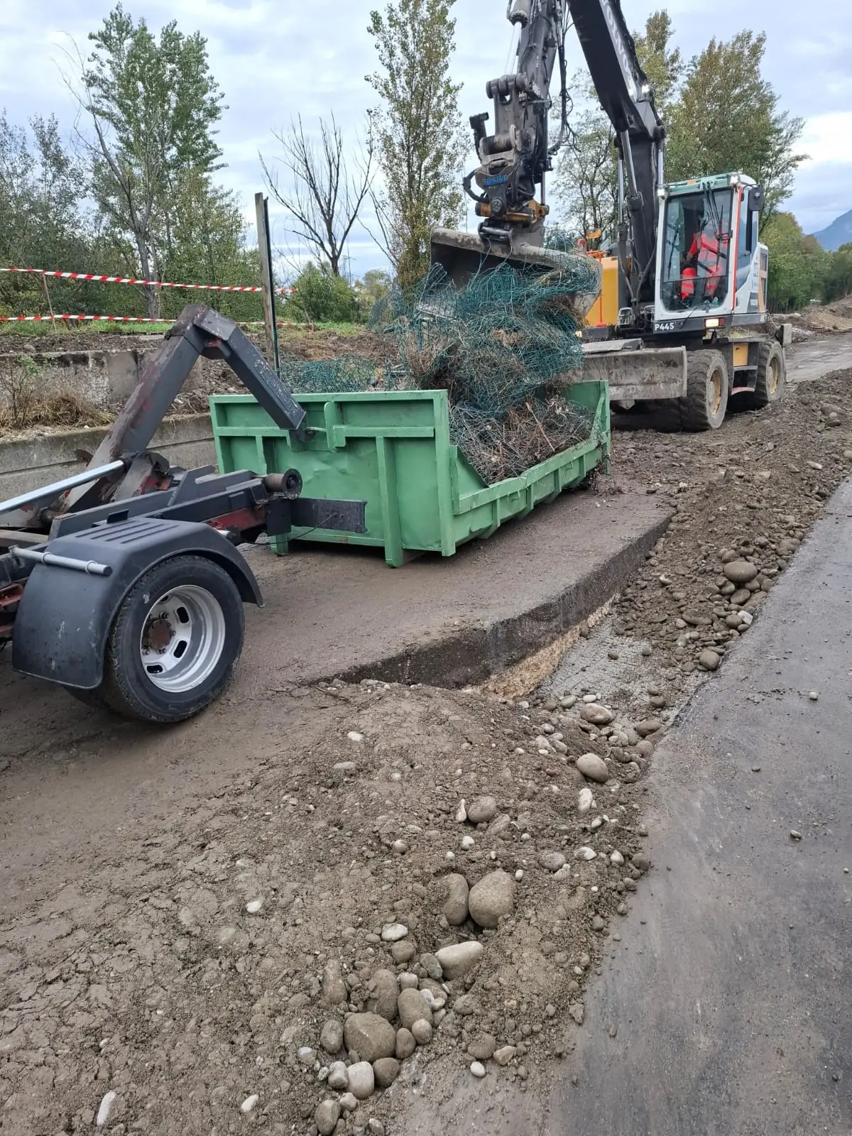 Une benne verte posée au bord d’un chantier de voirie : une pelle mécanique y déverse du grillage arraché ; à gauche, le bras à crochet qui pose et reprend la benne.