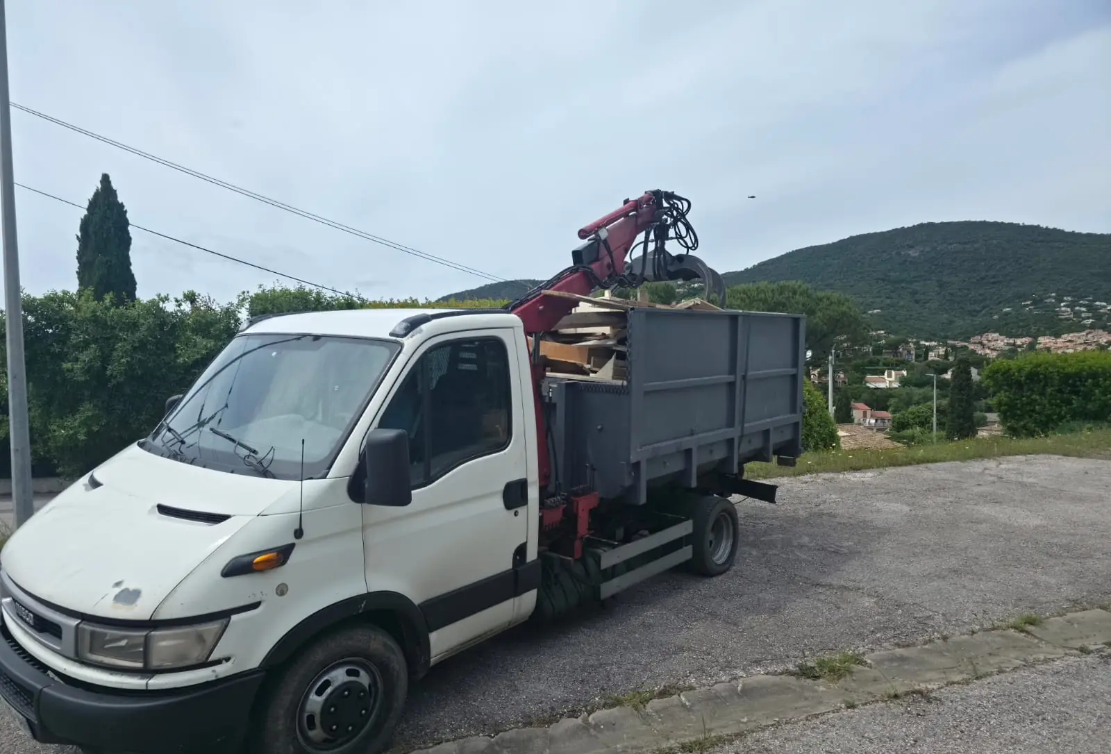 Le camion de RJ Benne garé au bord d’une route, sa benne grise chargée de planches et de bois de démolition, le grappin levé au-dessus.